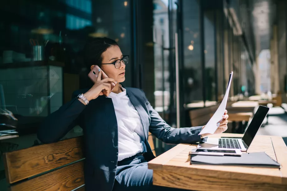 Calm successful trendy woman talking on mobile phone while reading corporate documents during remote job on laptop in cafe patio