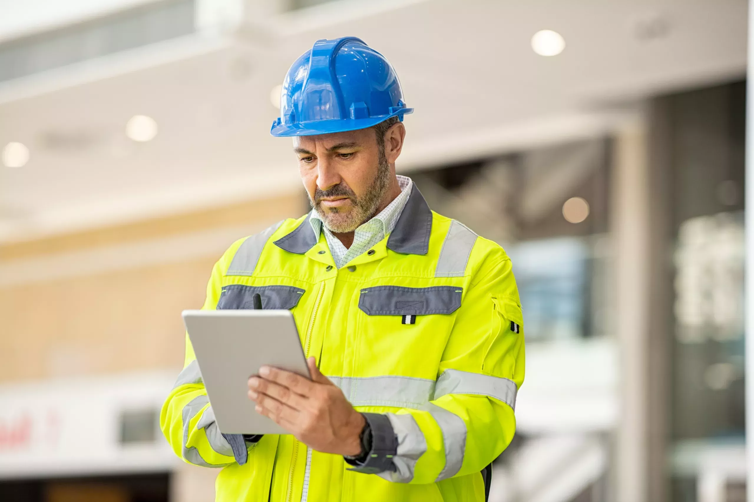 Serious and focused architect or engineer working on construction site using digital tablet while wearing safety vest and helmet. Mature man working on digital tablet at construction site. Mid adult manual worker with blue hardhat inspecting construction site while checking list on laptop.