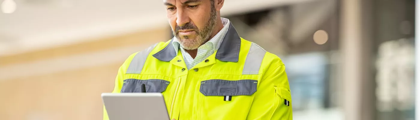 Serious and focused architect or engineer working on construction site using digital tablet while wearing safety vest and helmet. Mature man working on digital tablet at construction site. Mid adult manual worker with blue hardhat inspecting construction site while checking list on laptop.