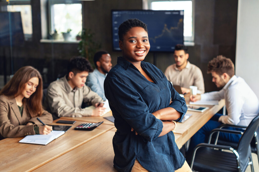 Modern entrepreneur concept. African american businesswoman standing with folded arms in conference room during meeting with colleagues and smiling at camera
